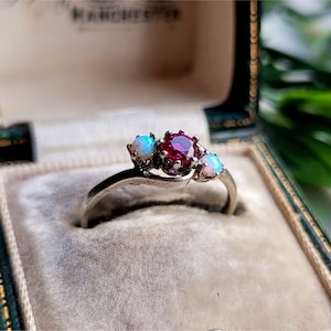 Op de afbeelding: Een zilveren ring met een roze edelsteen en twee witte opalen. De ring ligt in een groene fluwelen ringdoos.