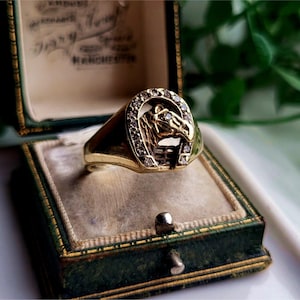 Puede incluir: Un anillo de oro con un diseño de herradura engastado con diamantes. La herradura está adornada con una cabeza de caballo.