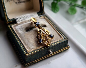 May include: A gold pendant with a leaf design, featuring small diamonds and dark blue gemstones. The pendant is displayed in a vintage green jewelry box with gold trim and a soft, light-colored interior.