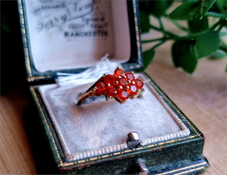 May include: A gold ring with a cluster of red gemstones set in a flower shape. The ring is in a green velvet ring box.