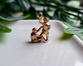 Könnte beinhalten: Goldener Anhänger mit zwei Figuren, eine hält einen Ring, die andere hält die Hand der ersten Figur. Die Figuren stehen auf einer weißen Oberfläche.