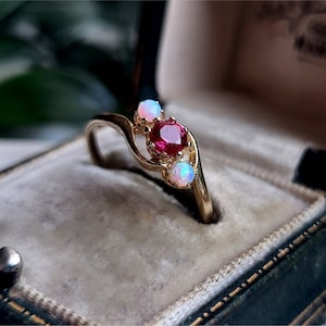 May include: A gold ring with a swirling design, featuring a central red gemstone flanked by two smaller opalescent stones. The ring is displayed in a vintage jewelry box with a velvet interior.