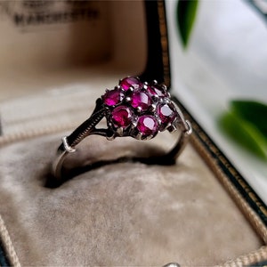 May include: A silver ring featuring a cluster of deep red gemstones set in a delicate floral design. The ring is displayed in a velvet-lined jewellery box.