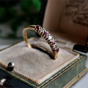 Puede incluir: Un anillo de banda de oro con rubíes rojos y diamantes blancos alternados. El anillo está en una caja de anillo de terciopelo verde.