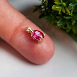 May include: A small oval-cut vibrant pink gemstone pendant set in a gold-toned metal prong setting. The pendant features a polished gold bail at the top for attachment to a chain, showcasing a faceted surface that reflects light across the stone.