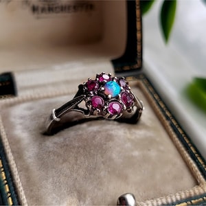 Könnte beinhalten: Ein silberner Ring mit einem großen Opal-Edelstein, der von kleineren Rubinen umgeben ist. Der Ring befindet sich in einer samtenen Schmuckschatulle.