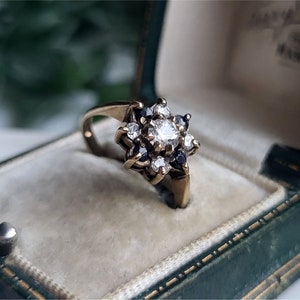 Puede incluir: Un anillo de oro con un racimo de pequeños diamantes y zafiros en forma de flor. El anillo está en una caja de anillo de terciopelo verde.