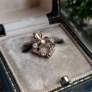 Könnte beinhalten: Ein goldener herzförmiger Anhänger mit roten und weißen Edelsteinen. Der Anhänger befindet sich in einer grünen Samt-Schmuckschatulle.