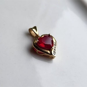 May include: A gold heart-shaped pendant featuring a large red gemstone in the centre and smaller clear gemstones around the edge.