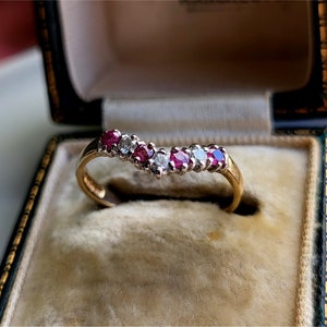 May include: A gold band ring with a curved row of red and white gemstones. The ring is in a velvet-lined box.