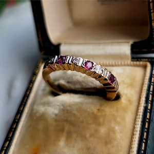 Può includere: Un anello a fascia d'oro con pietre preziose rosse e bianche alternate. L'anello è in una scatola foderata in velluto.