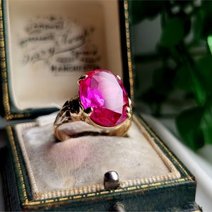 Könnte beinhalten: Ein goldener Ring mit einem großen, ovalen, leuchtend pinkfarbenen Edelstein. Der Ring befindet sich in einer grünen Samt-Ringbox.
