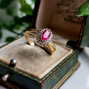 May include: A gold ring featuring a large oval ruby surrounded by smaller diamonds. The ring is displayed in a green velvet ring box.