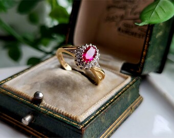 Pode incluir: Um anel de ouro com uma grande rubi ovalada cercada por diamantes menores. O anel está em uma caixa de anel de veludo verde.