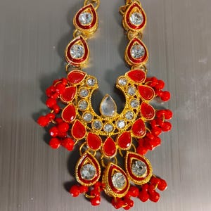 May include: A gold-tone necklace featuring a central crescent-shaped pendant adorned with red teardrop-shaped stones and clear crystals. The design includes dangling red beads and a chain with matching red and crystal teardrop accents.