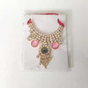 Peut inclure: Un collier décoratif composé de strass transparents sertis dans du métal doré, agrémenté de deux cabochons roses ronds et d'un motif central en plume de paon. Le bijou comprend un cordon en tissu rouge et est présenté sur une carte blanche.