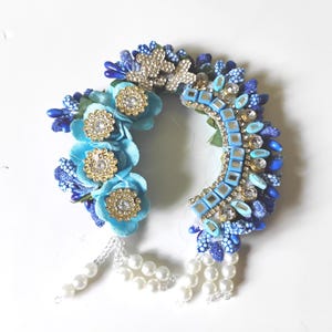 May include: A crescent-shaped hair accessory featuring light blue fabric flowers with gold-tone rhinestone centres, blue beads, clear crystals, and square mirror accents. The piece is finished with dangling white faux pearl bead strands.