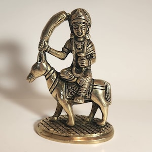 May include: A brass figurine depicting a deity riding a donkey, holding a curved object overhead and a small vessel. The figure is detailed with intricate clothing and jewelry. The base is round with a textured pattern.