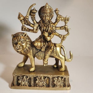 May include: A gold-coloured metal statue of a Hindu deity, Durga, riding a lion. She has eight arms and is holding various weapons and symbols. The statue is on a square base with intricate carvings.