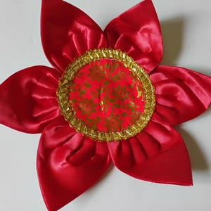 Könnte beinhalten: Eine sechsblättrige Stoffblume als Dekoration mit einer leuchtend roten Satin-Oberfläche. Die Mitte besteht aus einem runden Stoffeinsatz in Rot mit goldenem Blumenmuster, umrandet von einer dekorativen, strukturierten Goldborte.