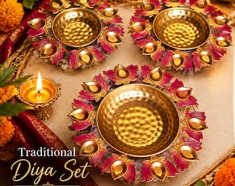 May include: A set of three round metal diya oil lamps featuring a central hammered gold bowl surrounded by pink lotus petal accents. One small lit diya lamp sits nearby. The text reads Traditional Diya Set LIGHT UP YOUR FESTIVITIES.
