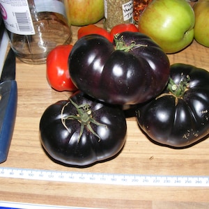 May include: A selection of dark purple heirloom tomatoes with green stems, red peppers, and green apples. A blue-handled knife and a ruler are on a wooden surface. The ruler shows measurements in centimetres.