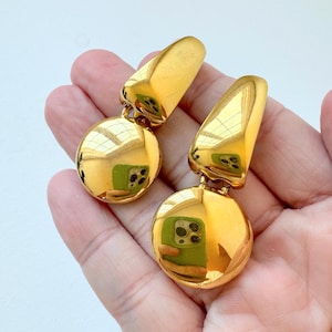 May include: A pair of gold-colored earrings. Each earring features a rounded, disc-shaped bottom and a triangular top. The earrings are highly reflective, with the light reflecting off the surface.