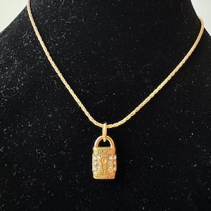 May include: Gold-toned necklace with a pendant shaped like a padlock. The pendant is encrusted with clear crystals and features the brand name "GIVENCHY" and "PARIS". The chain has a rope-like design, and the necklace is displayed against a black background.