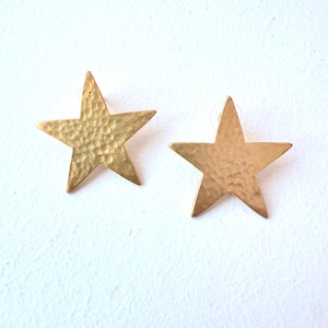 May include: Two gold-colored star-shaped earrings. The earrings have a hammered texture and are set against a white background. The star earrings are a celestial-themed accessory.