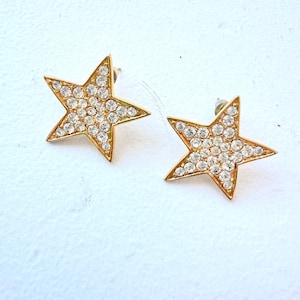May include: A pair of gold-toned star-shaped stud earrings. Each earring is encrusted with numerous clear, sparkling rhinestones. The earrings are set against a white background, highlighting their design and details.