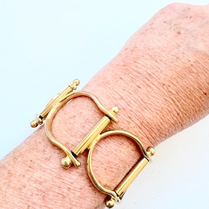 May include: A gold-toned metal cuff bracelet featuring a double stirrup-inspired design. The piece consists of two rounded, open-loop shapes connected by a central horizontal bar, with small spherical finials at each end of the metal frame.