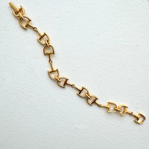 May include: A gold-toned metal bracelet composed of a series of repeating horse bit links. The chain features a polished finish and a secure clasp mechanism at one end, creating a continuous, articulated design of connected equestrian-inspired hardware.