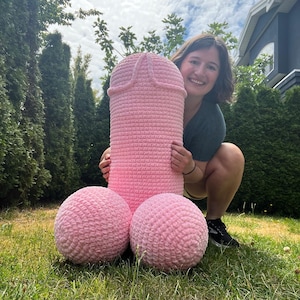 May include: A large pink crocheted sculpture resembling a human male anatomy. The sculpture is sitting on green grass with a person crouching behind it.