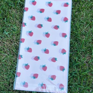 K&ouml;nnte beinhalten: Ein rechteckiges Notizbuch mit einem sich wiederholenden Muster aus roten Erdbeeren mit gr&uuml;nen Stielen und blauen Schatten auf hellrosa Hintergrund. Das Notizbuch ist in klarem Kunststoff verpackt.