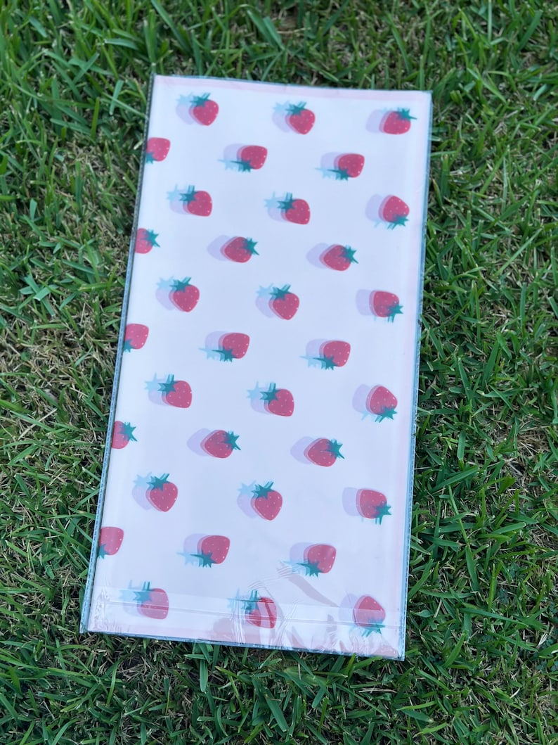 K&ouml;nnte beinhalten: Ein rechteckiges Notizbuch mit einem sich wiederholenden Muster aus roten Erdbeeren mit gr&uuml;nen Stielen und blauen Schatten auf hellrosa Hintergrund. Das Notizbuch ist in klarem Kunststoff verpackt.