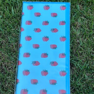 K&ouml;nnte beinhalten: Eine rechteckige, hellblaue Notizbuchh&uuml;lle mit einem sich wiederholenden Muster aus roten Erdbeeren. Die Erdbeeren haben einen Doppeleffekt, und die H&uuml;lle ist in klarem Kunststoff eingeschlossen. Der Hintergrund ist gr&uuml;nes Gras.