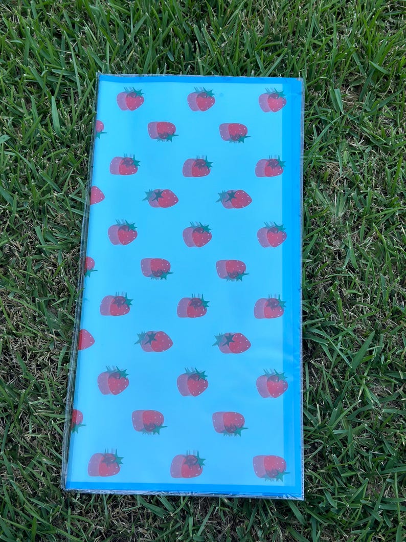 K&ouml;nnte beinhalten: Eine rechteckige, hellblaue Notizbuchh&uuml;lle mit einem sich wiederholenden Muster aus roten Erdbeeren. Die Erdbeeren haben einen Doppeleffekt, und die H&uuml;lle ist in klarem Kunststoff eingeschlossen. Der Hintergrund ist gr&uuml;nes Gras.