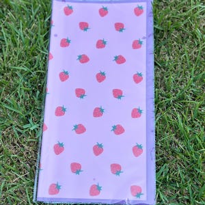 K&ouml;nnte beinhalten: Eine transparente Kunststoffh&uuml;lle mit einem lavendelfarbenen Blatt mit einem sich wiederholenden Muster aus rosa Erdbeeren mit gr&uuml;nen Stielen. Die H&uuml;lle ist rechteckig und scheint eine Notizbuchh&uuml;lle oder ein Planereinsatz zu sein.