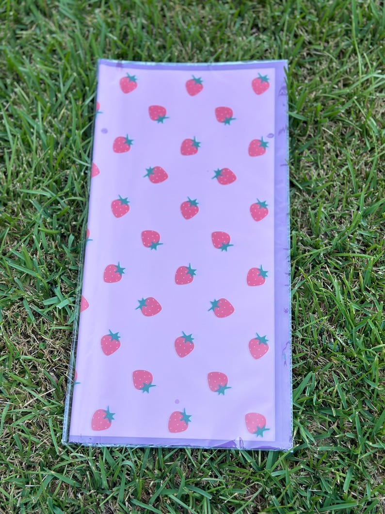 K&ouml;nnte beinhalten: Eine transparente Kunststoffh&uuml;lle mit einem lavendelfarbenen Blatt mit einem sich wiederholenden Muster aus rosa Erdbeeren mit gr&uuml;nen Stielen. Die H&uuml;lle ist rechteckig und scheint eine Notizbuchh&uuml;lle oder ein Planereinsatz zu sein.