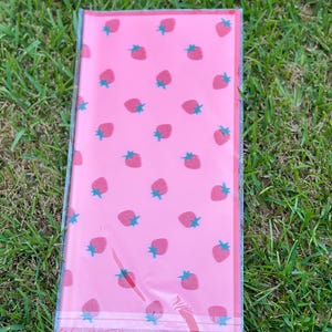 K&ouml;nnte beinhalten: Ein rechteckiges, rosa Paket, verpackt in durchsichtigem Kunststoff. Das Paket ist mit einem Muster aus roten Erdbeeren mit gr&uuml;nen Stielen versehen. Das Paket liegt auf gr&uuml;nem Gras.