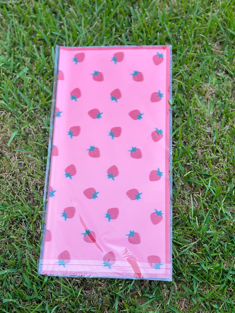 K&ouml;nnte beinhalten: Ein rechteckiges, rosa Paket, verpackt in durchsichtigem Kunststoff. Das Paket ist mit einem Muster aus roten Erdbeeren mit gr&uuml;nen Stielen versehen. Das Paket liegt auf gr&uuml;nem Gras.