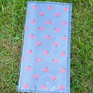 K&ouml;nnte beinhalten: Ein rechteckiges Notizbuch mit grauem Einband und einem Muster aus rosa Erdbeeren mit gr&uuml;nen Stielen. Das Notizbuch ist in klarem Kunststoff verpackt und liegt auf gr&uuml;nem Gras.