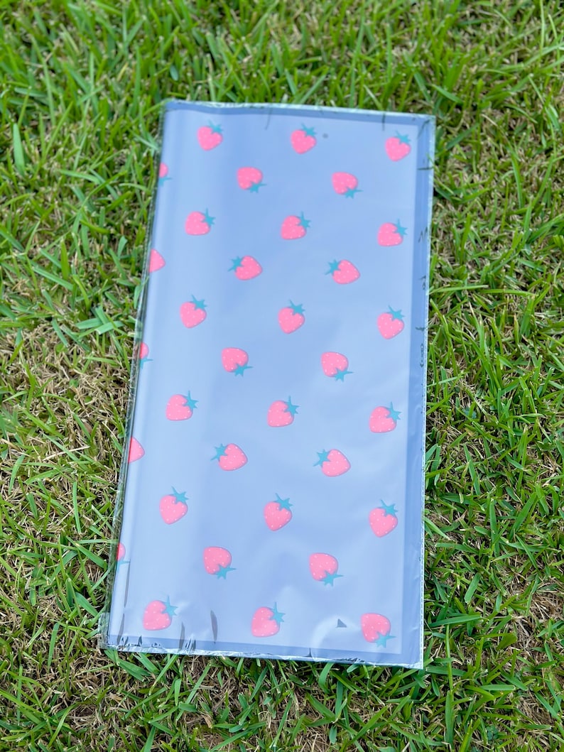 K&ouml;nnte beinhalten: Ein rechteckiges Notizbuch mit grauem Einband und einem Muster aus rosa Erdbeeren mit gr&uuml;nen Stielen. Das Notizbuch ist in klarem Kunststoff verpackt und liegt auf gr&uuml;nem Gras.