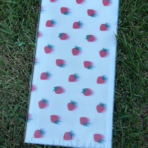 K&ouml;nnte beinhalten: Ein rechteckiges, klares Kunststoffpaket mit einem sich wiederholenden Muster aus roten Erdbeeren mit gr&uuml;nen Stielen auf wei&szlig;em Hintergrund. Das Paket liegt auf gr&uuml;nem Gras.