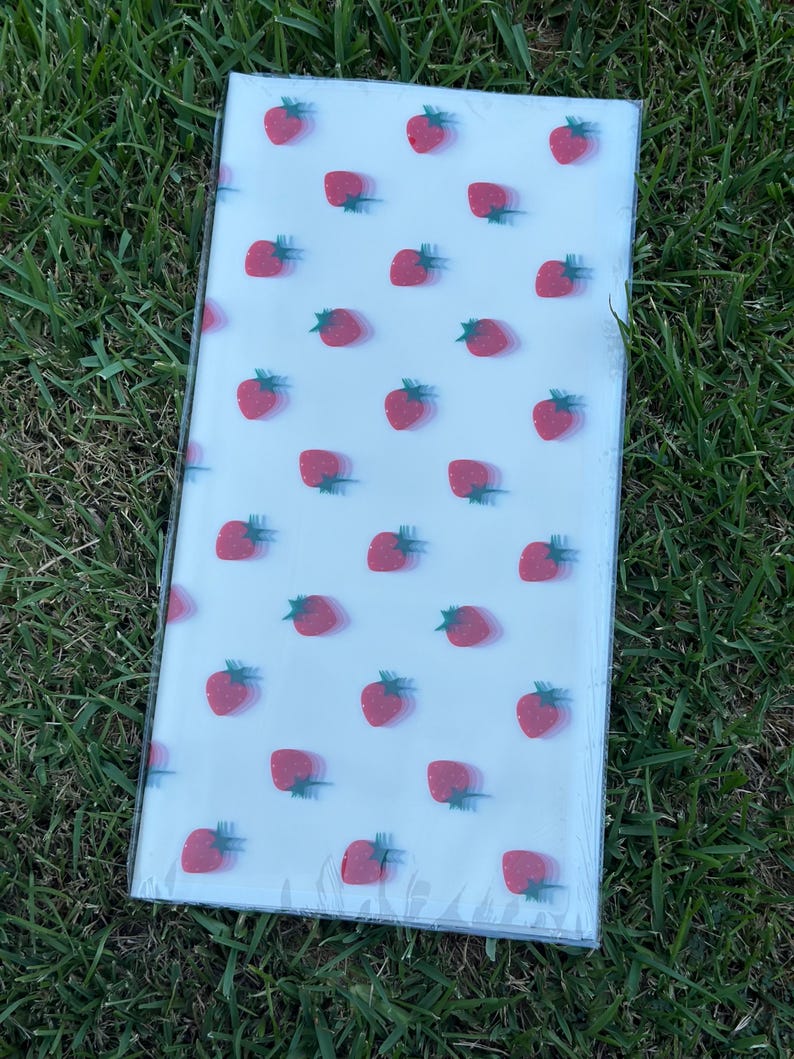 K&ouml;nnte beinhalten: Ein rechteckiges, klares Kunststoffpaket mit einem sich wiederholenden Muster aus roten Erdbeeren mit gr&uuml;nen Stielen auf wei&szlig;em Hintergrund. Das Paket liegt auf gr&uuml;nem Gras.