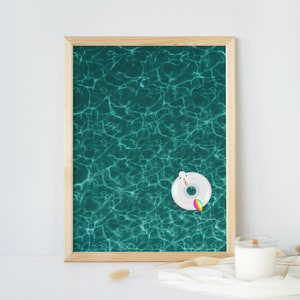 Può includere: Una stampa incorniciata raffigurante una superficie d'acqua turchese con riflessi di luce. Un galleggiante gonfiabile bianco a forma di unicorno con coda arcobaleno galleggia sull'acqua. La cornice è in legno chiaro.