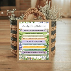 May include: A framed print with the text "Anxiety Coping Statements" and affirmations. The print has a wooden frame and is decorated with floral elements. It is displayed in a woven basket with dried plants.