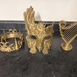 Può includere: Tre oggetti decorativi color oro: una corona, una maschera e un'arpa. La corona ha un design a punte. La maschera ha un design del viso ornato e dettagliato. L'arpa è su un piedistallo.