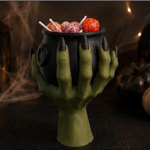 May include: A matte black cauldron bowl held by two green hands with long, pointed black fingernails. The cauldron is filled with several round, multi-colored lollipops on white sticks. The base of the object is a single, sturdy green pedestal.