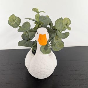 May include: A white ceramic goose planter with an orange beak, holding green leafy plants. The goose-shaped pot has a textured surface and is set against a black surface and white background. A decorative item for home decor.